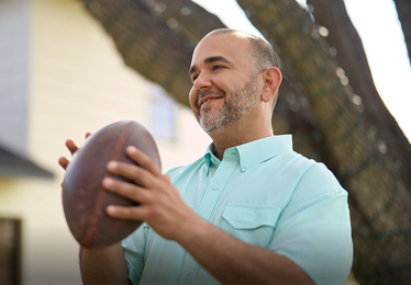 Hombre sonriendo mientras sostiene un balón de fútbol  americano al aire libre.